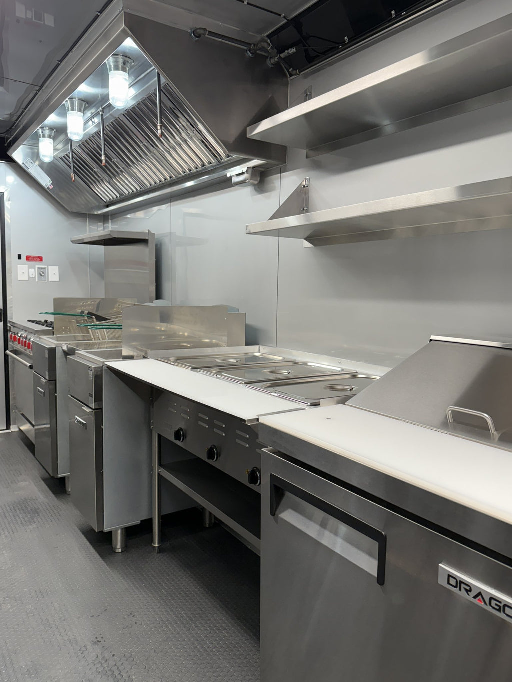 Commercial kitchen equipment including stainless steel ovens, fryers, a grill station, and a large under-counter refrigerator with a visible DRAGONE brand badge.
