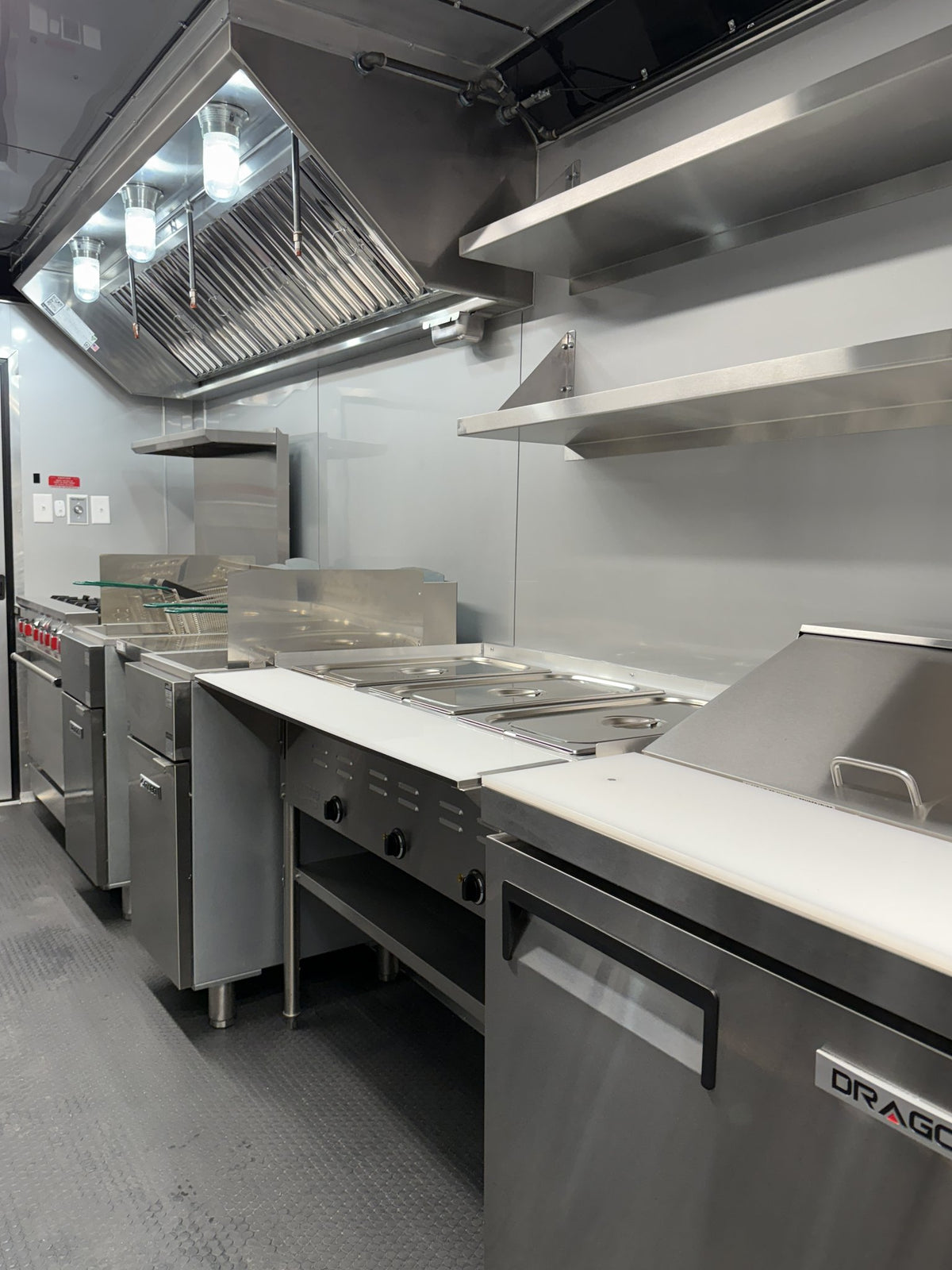 Commercial kitchen equipment including stainless steel ovens, fryers, a grill station, and a large under-counter refrigerator with a visible DRAGONE brand badge.