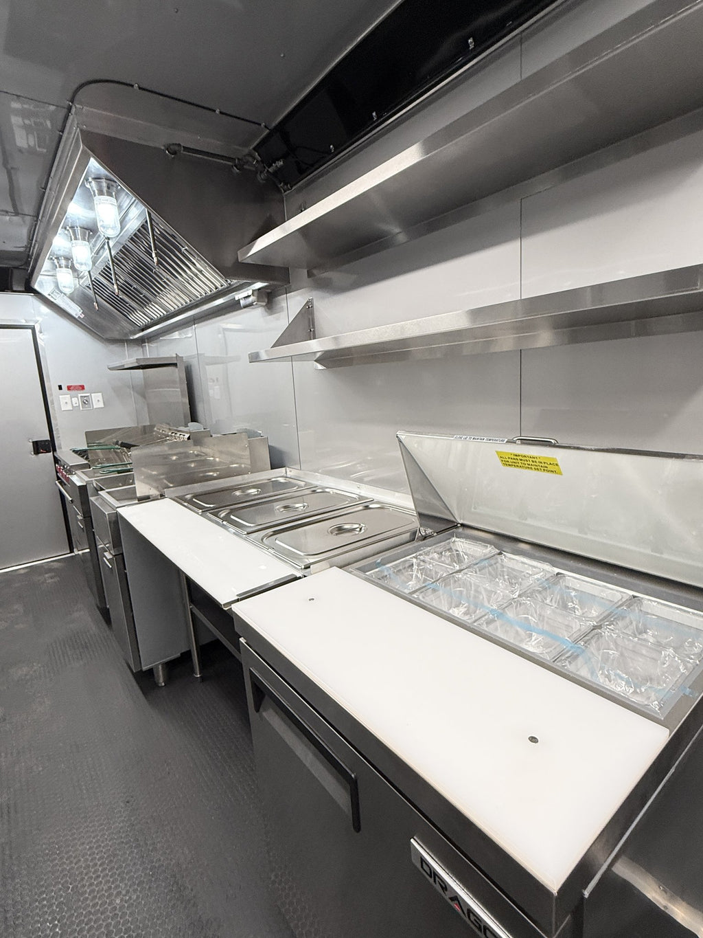 A commercial kitchen interior with stainless steel counters, food preparation areas, a large ventilation hood, shelving, and a gray textured floor.