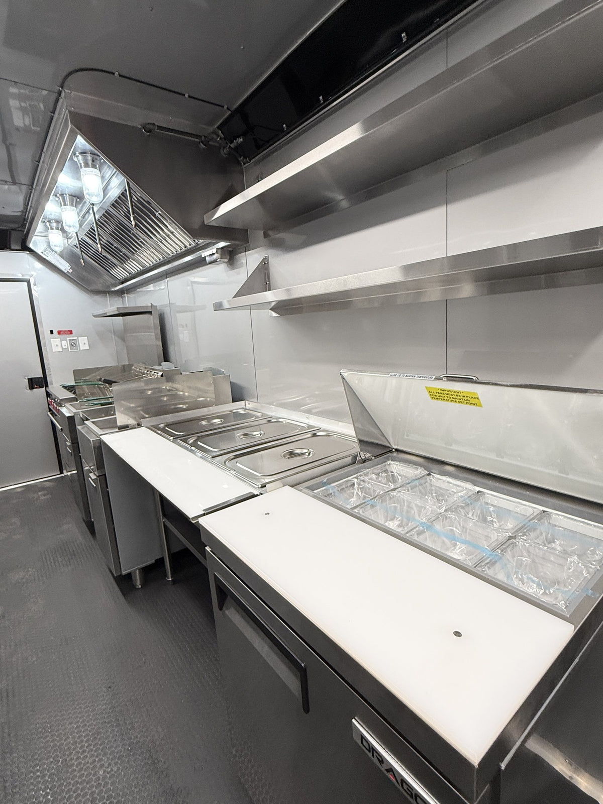 A commercial kitchen interior with stainless steel counters, food preparation areas, a large ventilation hood, shelving, and a gray textured floor.