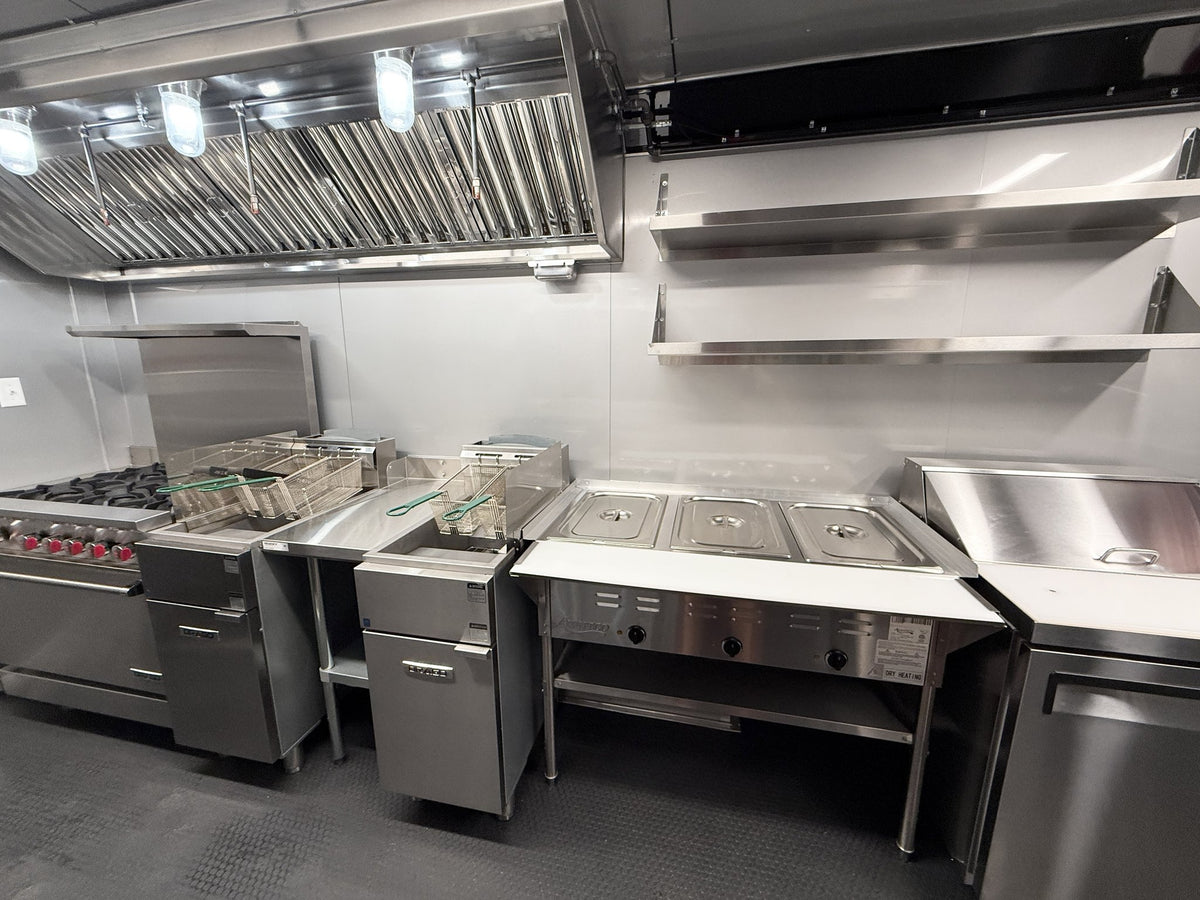 Commercial kitchen equipment including a stainless steel range, deep fryer, warming stations, and a large ventilation hood against a light gray tiled wall.