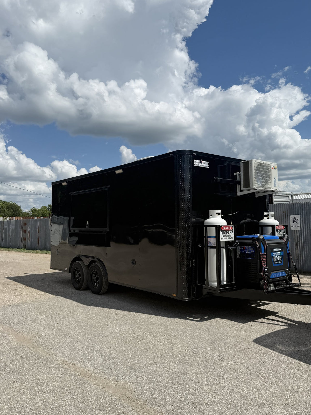 A black food truck trailer parked outdoors on a paved lot under a blue sky with large white clouds. The trailer has a side prep opening, a rear utility door, an air conditioner mounted on the upper right side, and gas cylinders and a blue generator attached at the back.