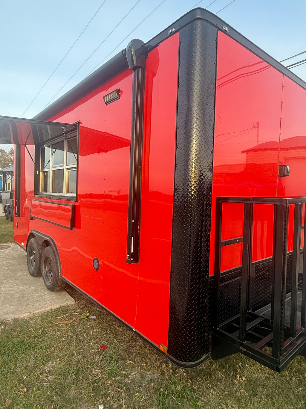 A bright red food truck trailer with black trim, a diamond-plate side panel, a service window, and a black metal ramp, photographed outdoors on grass and pavement.
