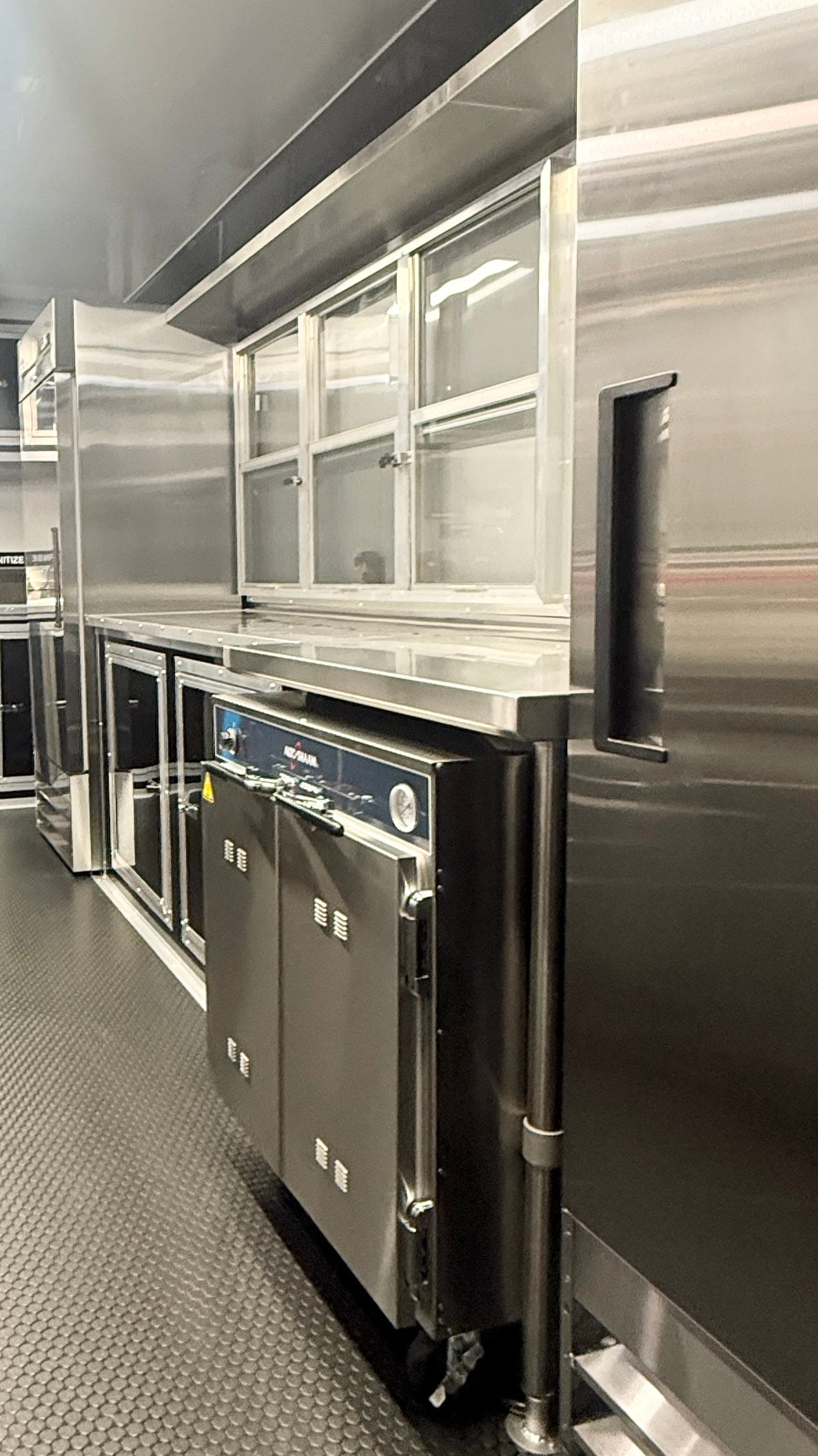 Commercial stainless steel kitchen equipment, including a double-door undercounter refrigerator or freezer, a prep table with glass-front upper cabinets, and a large stainless steel refrigerator or storage unit along the right side, set in a commercial kitchen environment.