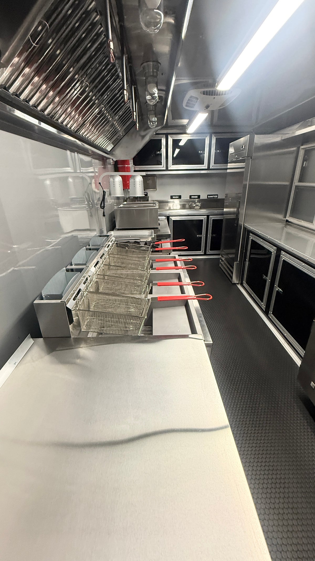 A stainless steel mobile kitchen interior with a long prep counter, hanging ventilation hood, built-in ovens, shelving, and a dark textured floor.