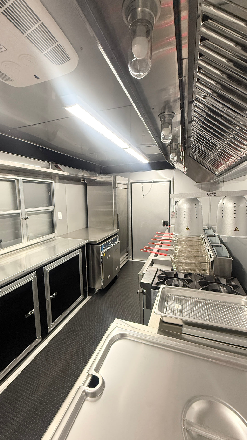 A commercial kitchen interior with stainless steel counters, prep tables, gas stoves, a large metal ventilation hood, refrigerated cabinets, and overhead fluorescent lighting.