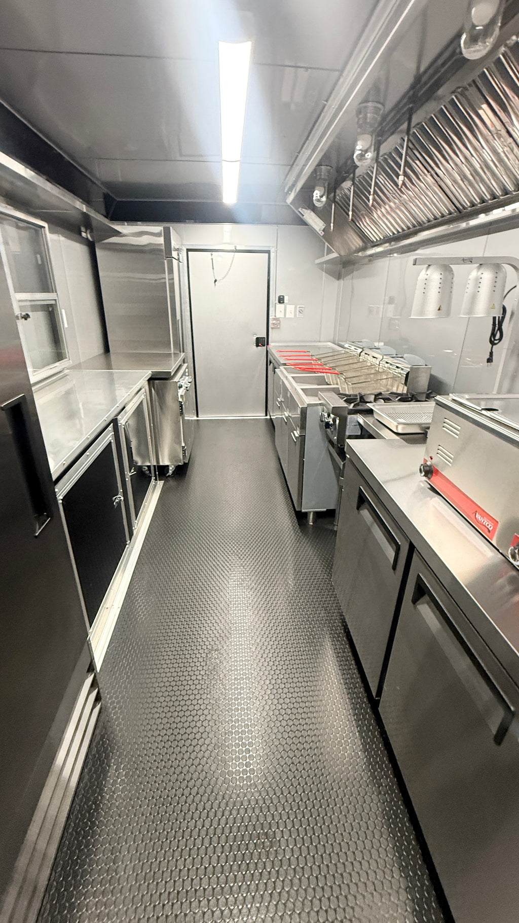 A commercial kitchen interior with stainless steel counters, prep tables, a large metal ventilation hood, a diamond-plate floor, and a white back wall.