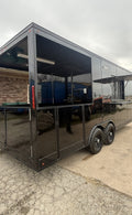 A black enclosed trailer or food truck with large side windows and a diamond plate frame, parked outdoors on a paved lot under an overcast sky.