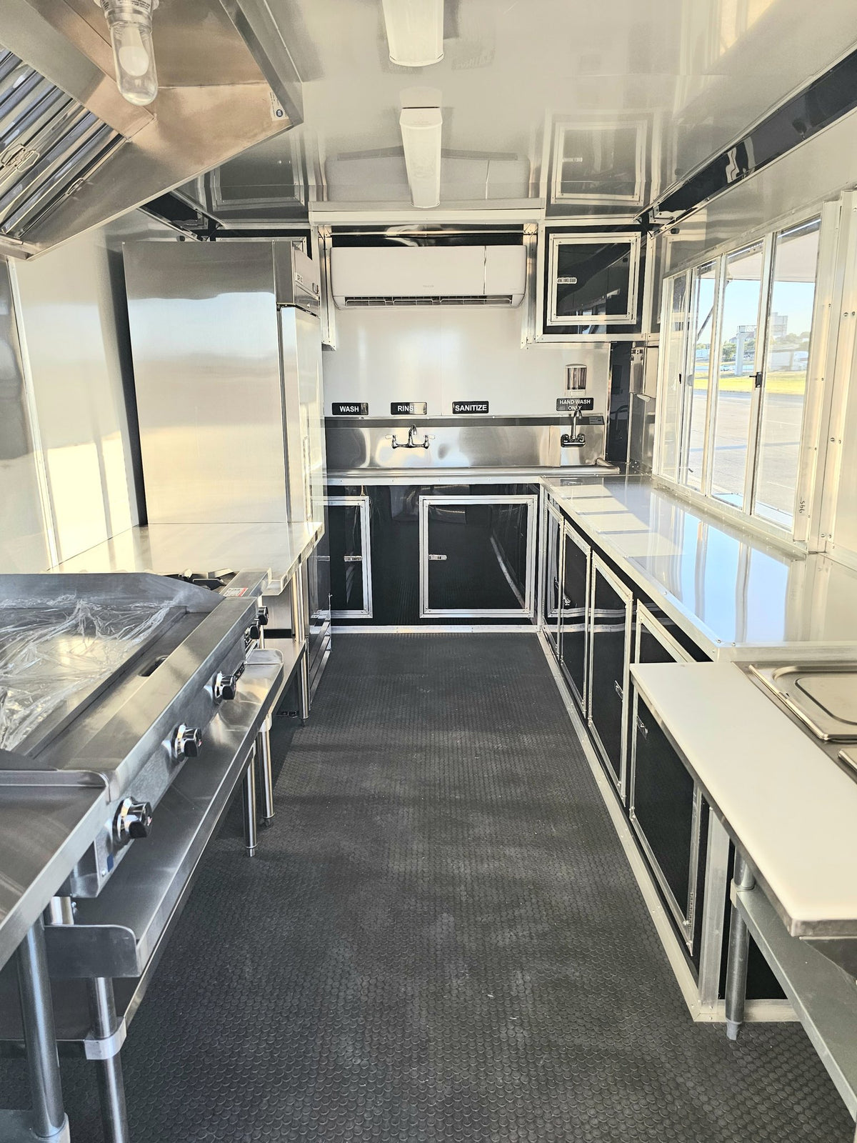 Commercial kitchen interior with stainless steel counters, appliances, and dark cabinetry in a food truck or kitchen trailer.