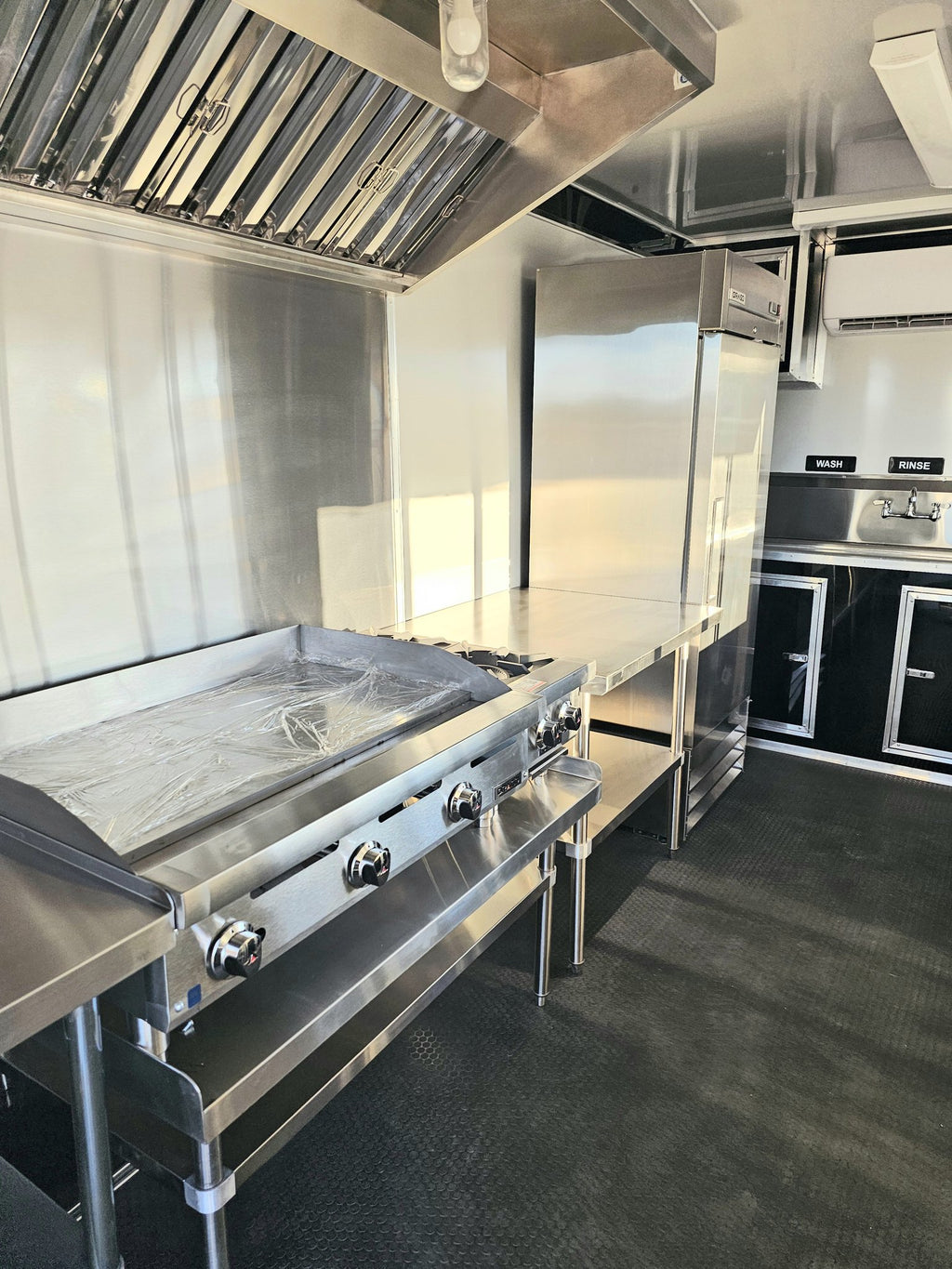 A commercial-grade stainless steel kitchen setup inside a trailer, featuring a large flat-top grill with a protective sheet, a tall refrigerator, a work surface, and dark cabinets with silver trim.