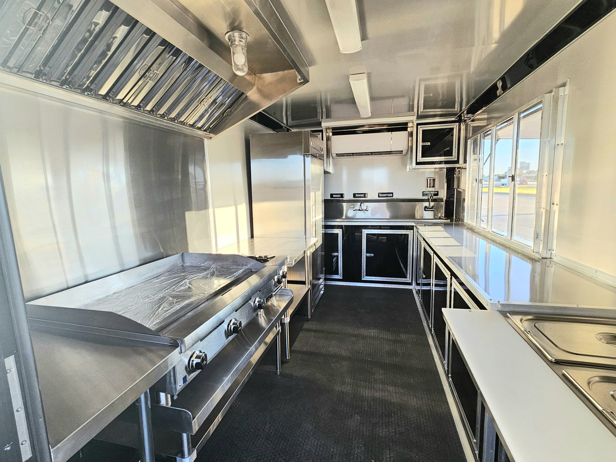 A stainless steel mobile kitchen interior with a large flat-top grill on the left, prep tables on the right, a sink area, and a window at the back.