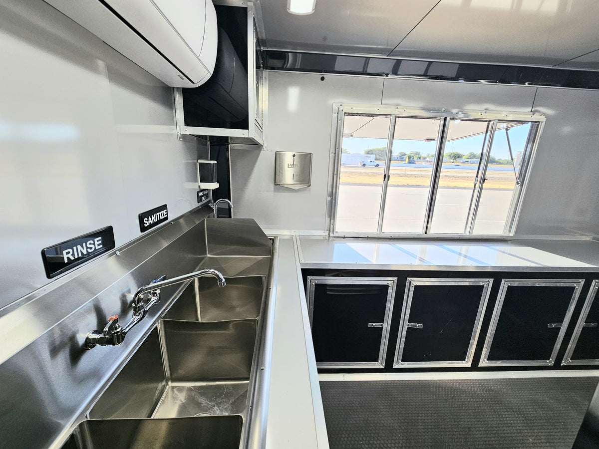 A clean, modern food truck kitchen interior featuring a stainless steel double sink with a chrome faucet, black and stainless steel cabinets, a white countertop, and a large window looking out onto a road.