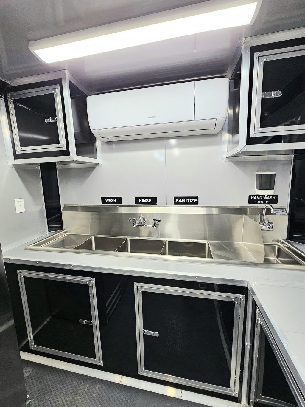 A compact kitchenette setup inside a vehicle or trailer, featuring stainless steel sinks, a backsplash, black and silver cabinets, a white air conditioner mounted on the wall, and labeled signs for wash, rinse, sanitize, and hand wash.