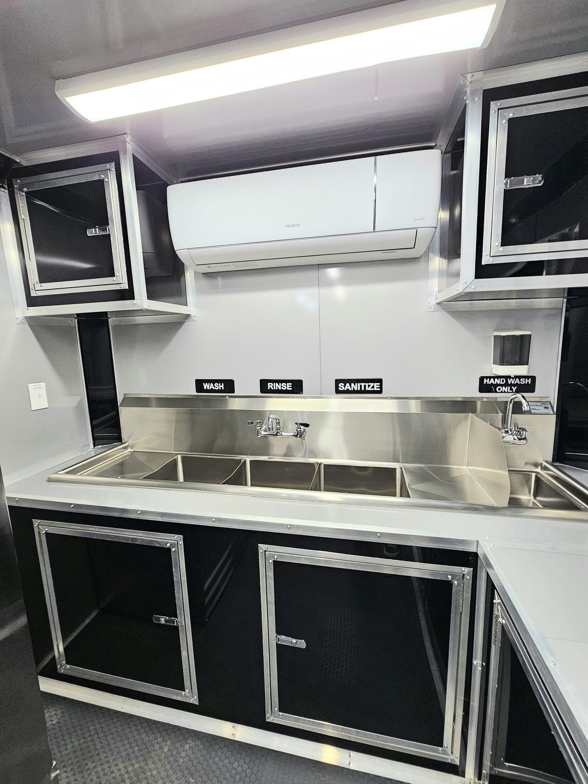 A compact kitchenette setup inside a vehicle or trailer, featuring stainless steel sinks, a backsplash, black and silver cabinets, a white air conditioner mounted on the wall, and labeled signs for wash, rinse, sanitize, and hand wash.