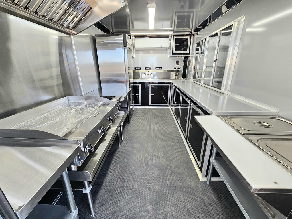 A commercial kitchen interior with stainless steel counters, prep tables, a large grill or smoker unit on the left, a sink area in the back, and a gray textured floor.