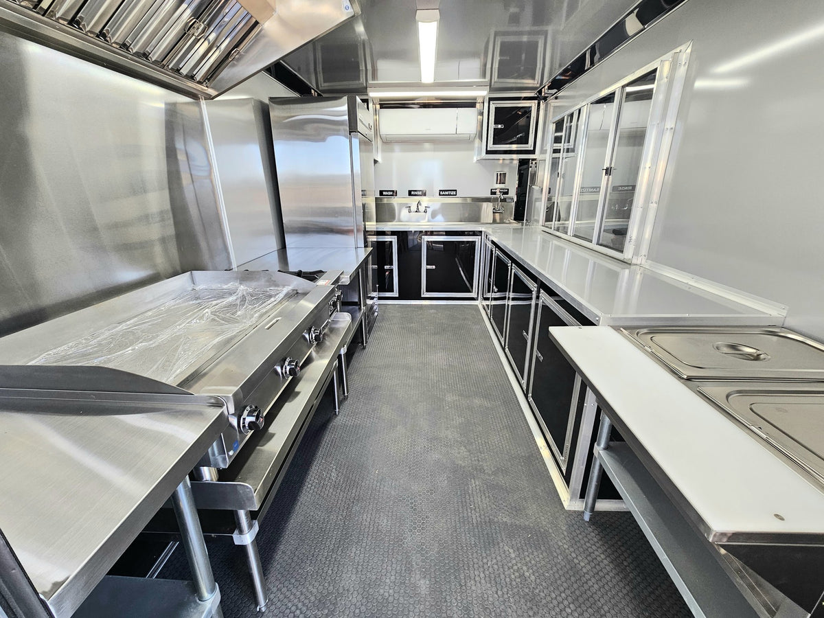 A commercial kitchen interior with stainless steel counters, prep tables, a large grill or smoker unit on the left, a sink area in the back, and a gray textured floor.