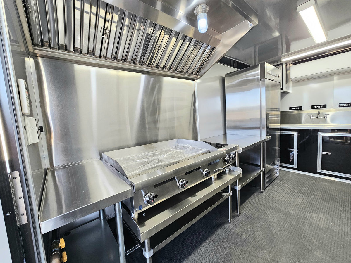 A commercial-grade stainless steel kitchen setup with a large flat-top grill, under-counter work surfaces, a tall refrigerator, and a ventilation hood in a clean, industrial kitchen environment.