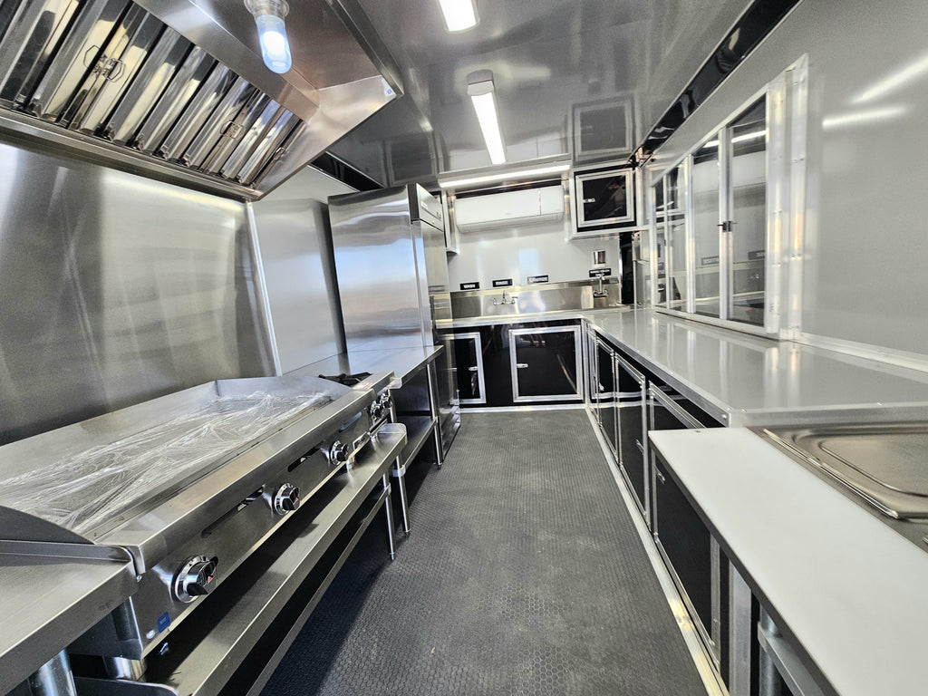 A commercial kitchen interior with stainless steel counters, a large grill or stove on the left, a refrigerator, sink area, and dark cabinetry along a narrow galley layout.