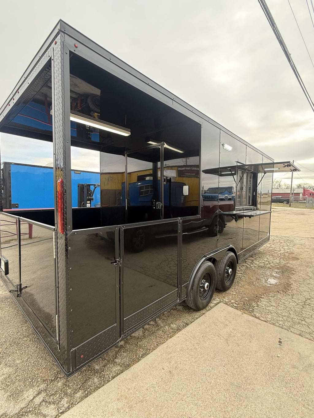 A black metal trailer with a large open side compartment and a small elevated service or food-service counter on the right, photographed outdoors on a concrete pad under an overcast sky.