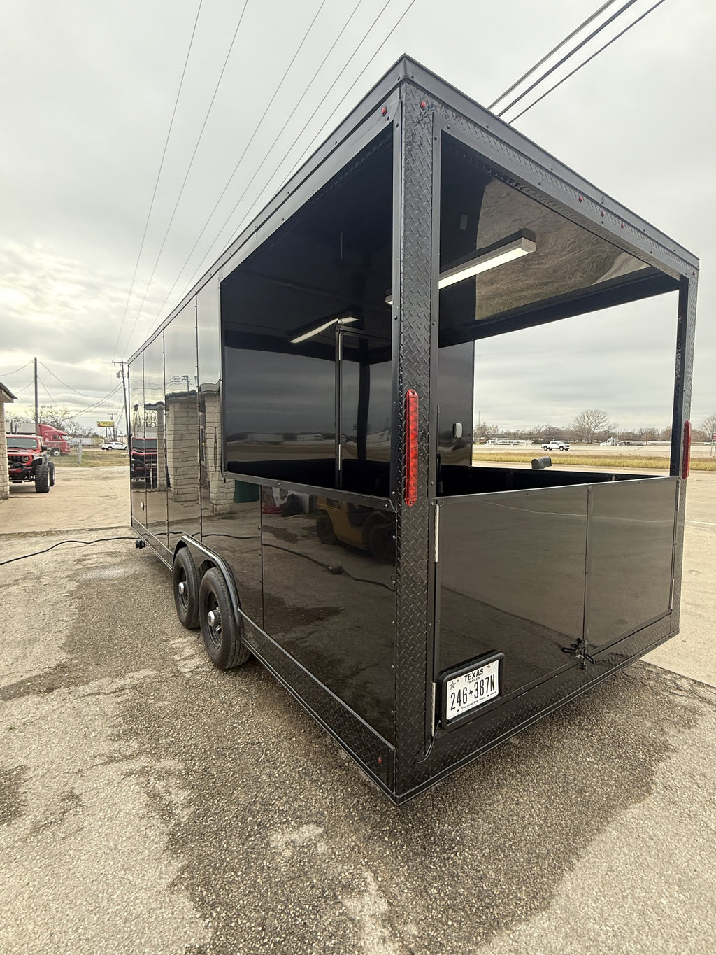 A black aluminum food truck trailer parked outdoors on a wet paved lot under an overcast sky. The trailer features a large open serving window on the side, a diamond plate texture on the front and side panels, dual rear wheels, and a license plate.