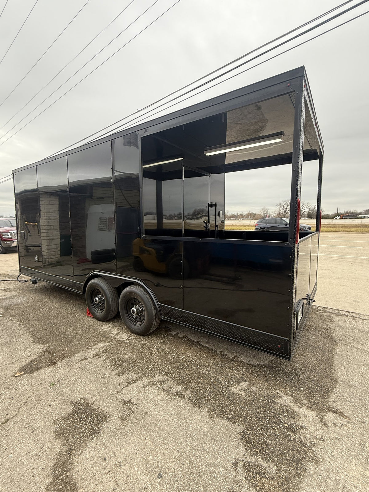 A black enclosed trailer with large glass windows and a side service door, parked outdoors on a wet paved lot under an overcast sky.