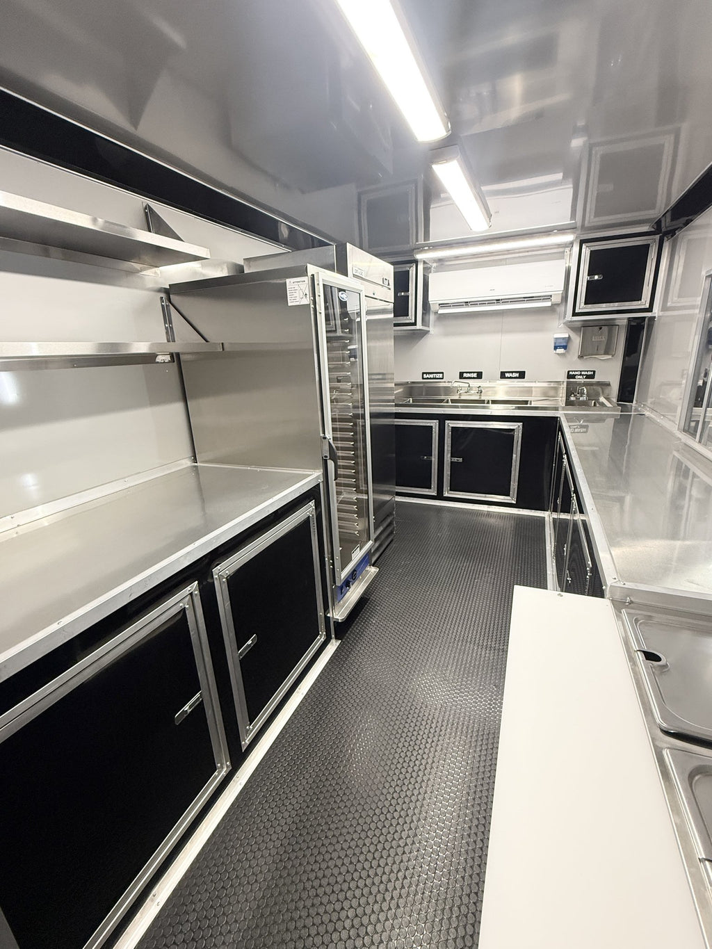 A commercial food prep kitchen interior with stainless steel counters, black cabinets, a tall glass-door refrigerator, sink area, and textured gray floor under bright overhead lights.