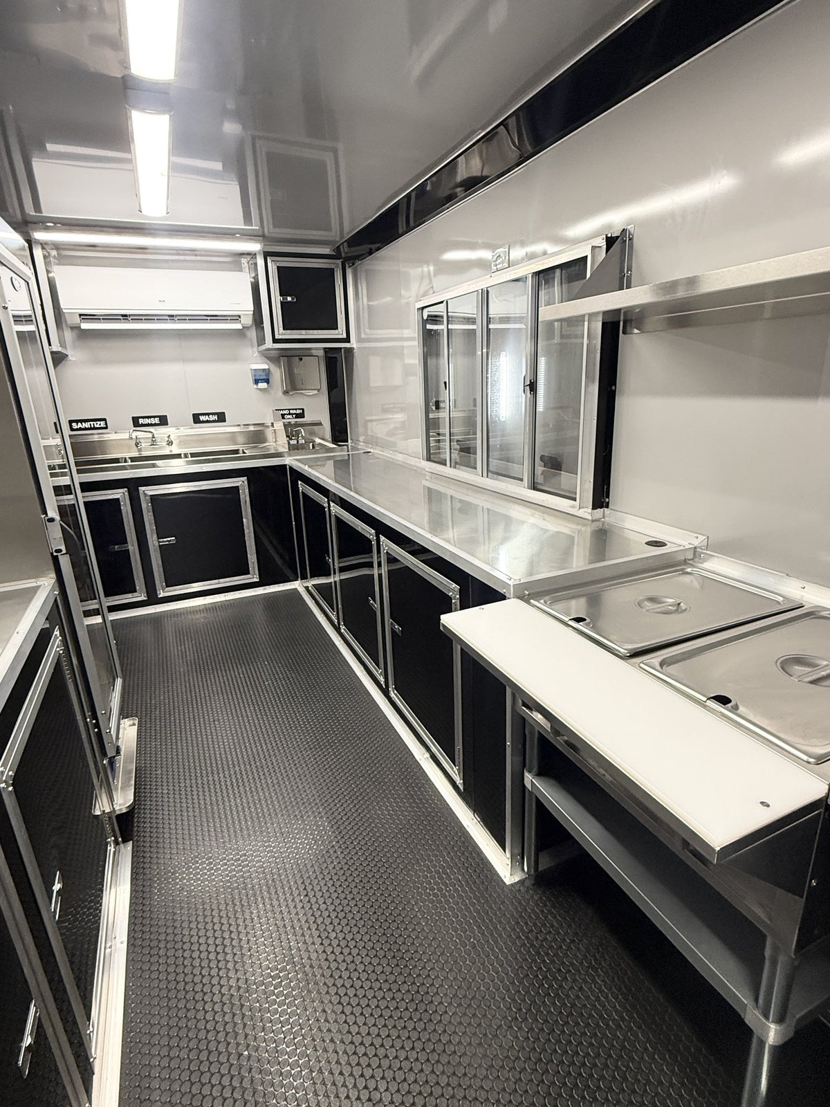 A stainless steel and black food truck kitchen interior with counters, prep tables, glass-front cabinets, and a textured dark floor.