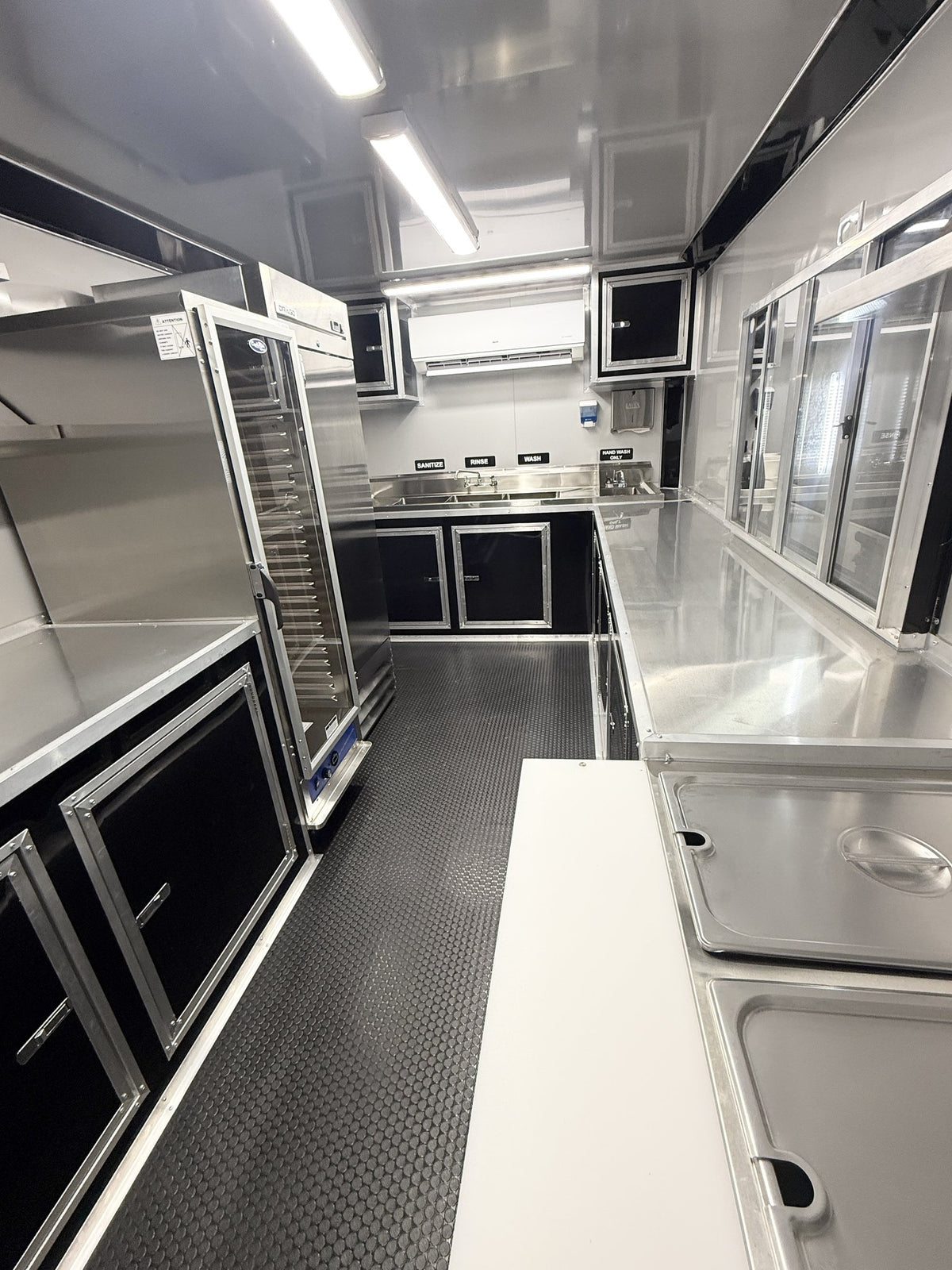 A commercial kitchen interior with stainless steel counters, glass-door fridges, wall-mounted cabinets, a textured dark floor, and bright overhead lighting.
