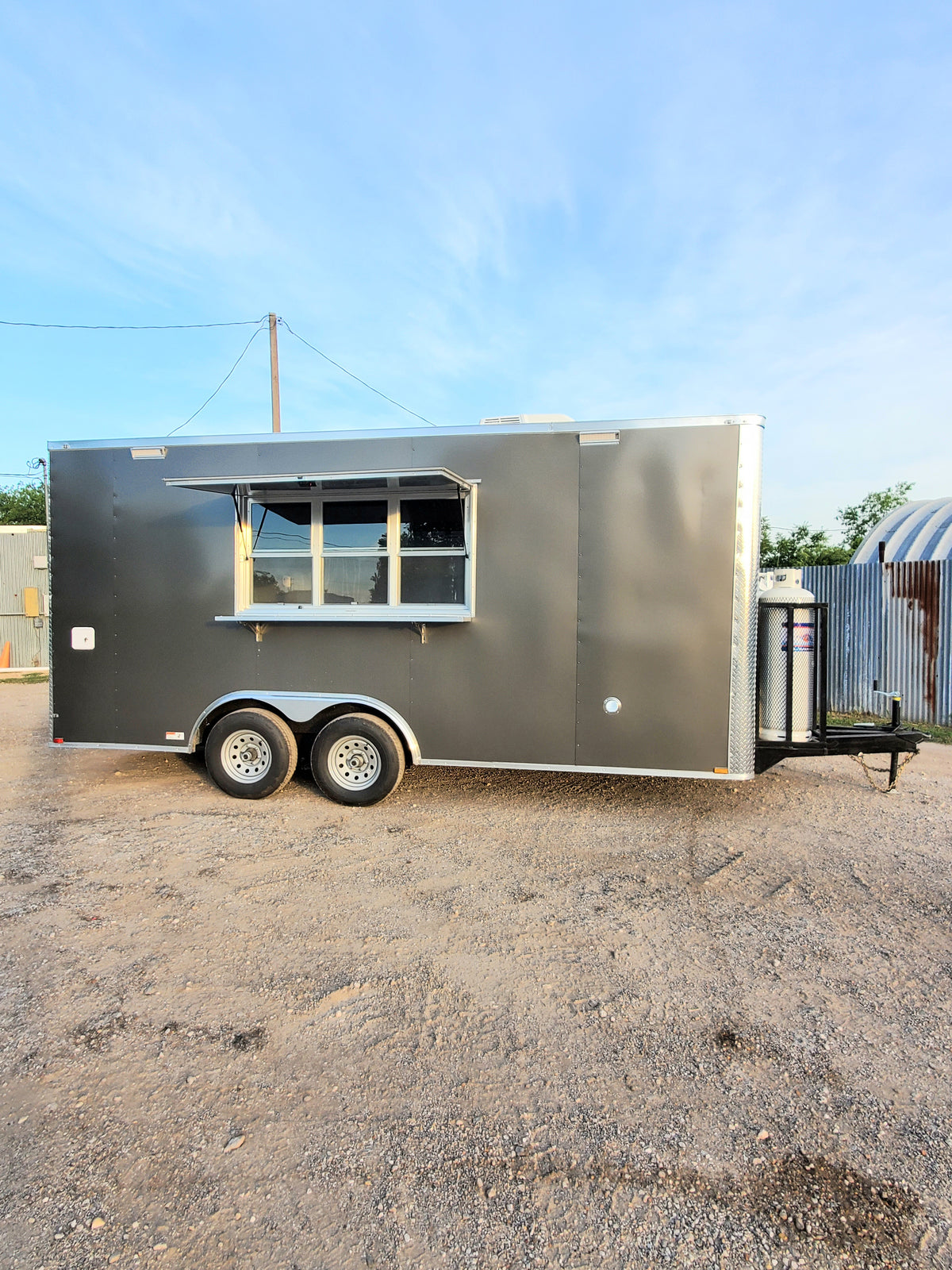 18 FT charcoal gray cooking trailer parked on gravel under a blue sky, side view with the serving window open