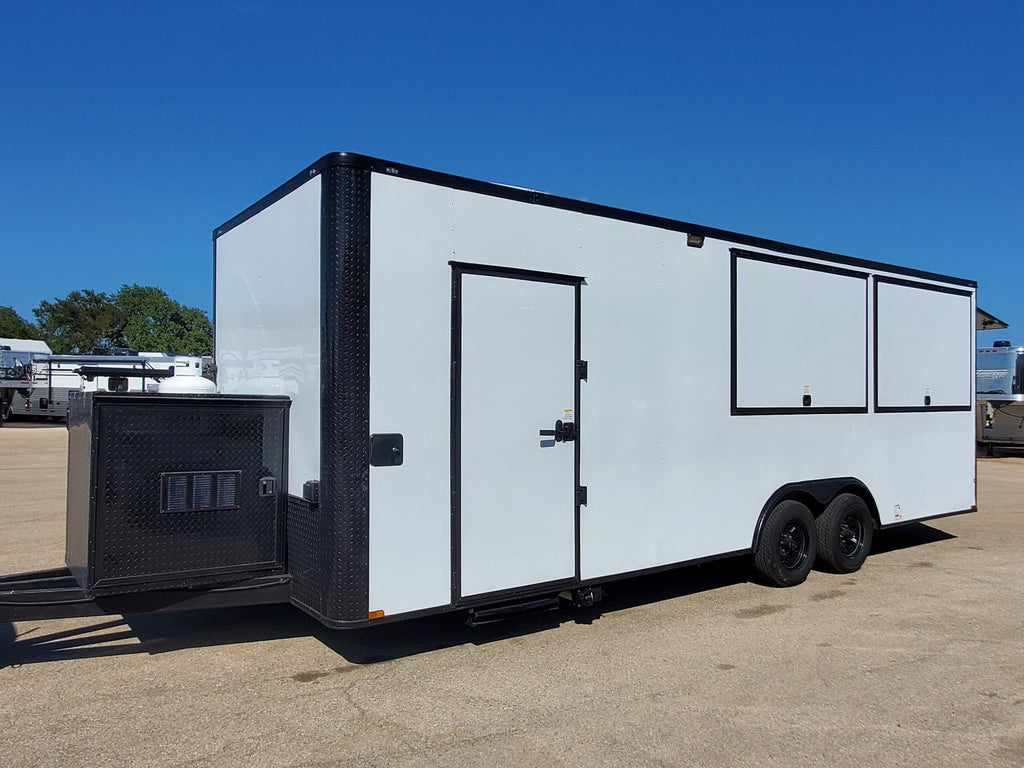 A white enclosed cargo trailer with black trim, a side door, and two large side windows, parked outdoors on a paved lot under a clear blue sky.