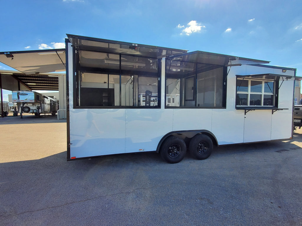 A white commercial food truck trailer with black trim, large windowed service windows, and dual rear wheels, photographed outdoors on a paved lot under a blue sky.