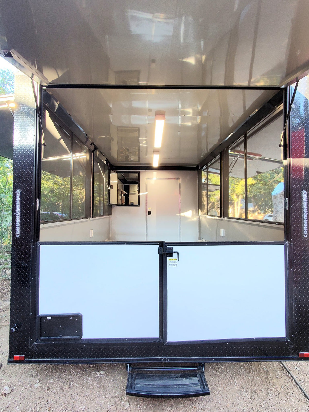 Rear view of a black enclosed trailer or cargo box with white side panels and a black step, photographed outdoors on a dirt ground with windows and interior lighting visible.