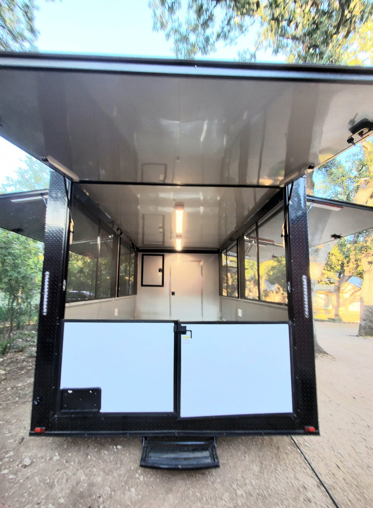 A black enclosed cargo trailer parked outdoors on a dirt surface with trees and a building visible in the background. The rear doors are open, showing a white interior with windows and a light fixture.