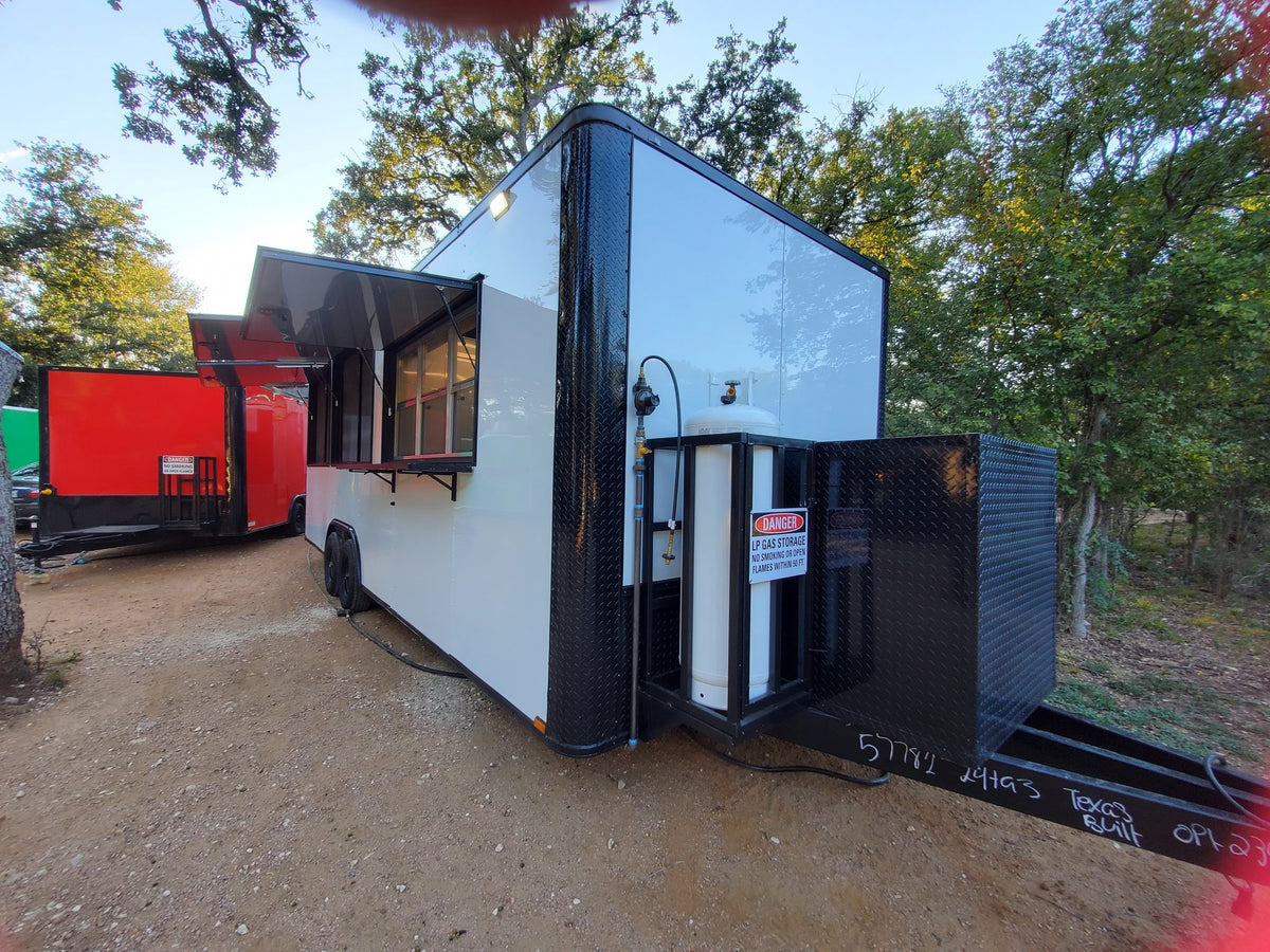 A white and black commercial food truck trailer parked outdoors on a dirt lot with trees in the background. The side window is open, and a black mechanical compartment or generator unit is extended from the rear side.