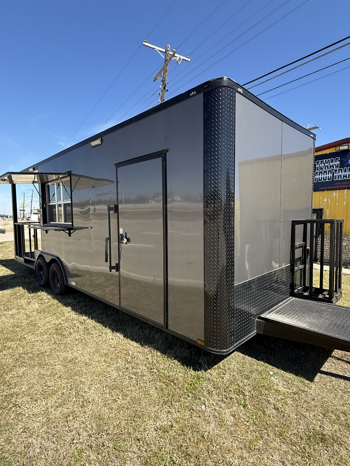 Black and gray enclosed trailer with a door on a grassy area under a clear blue sky.