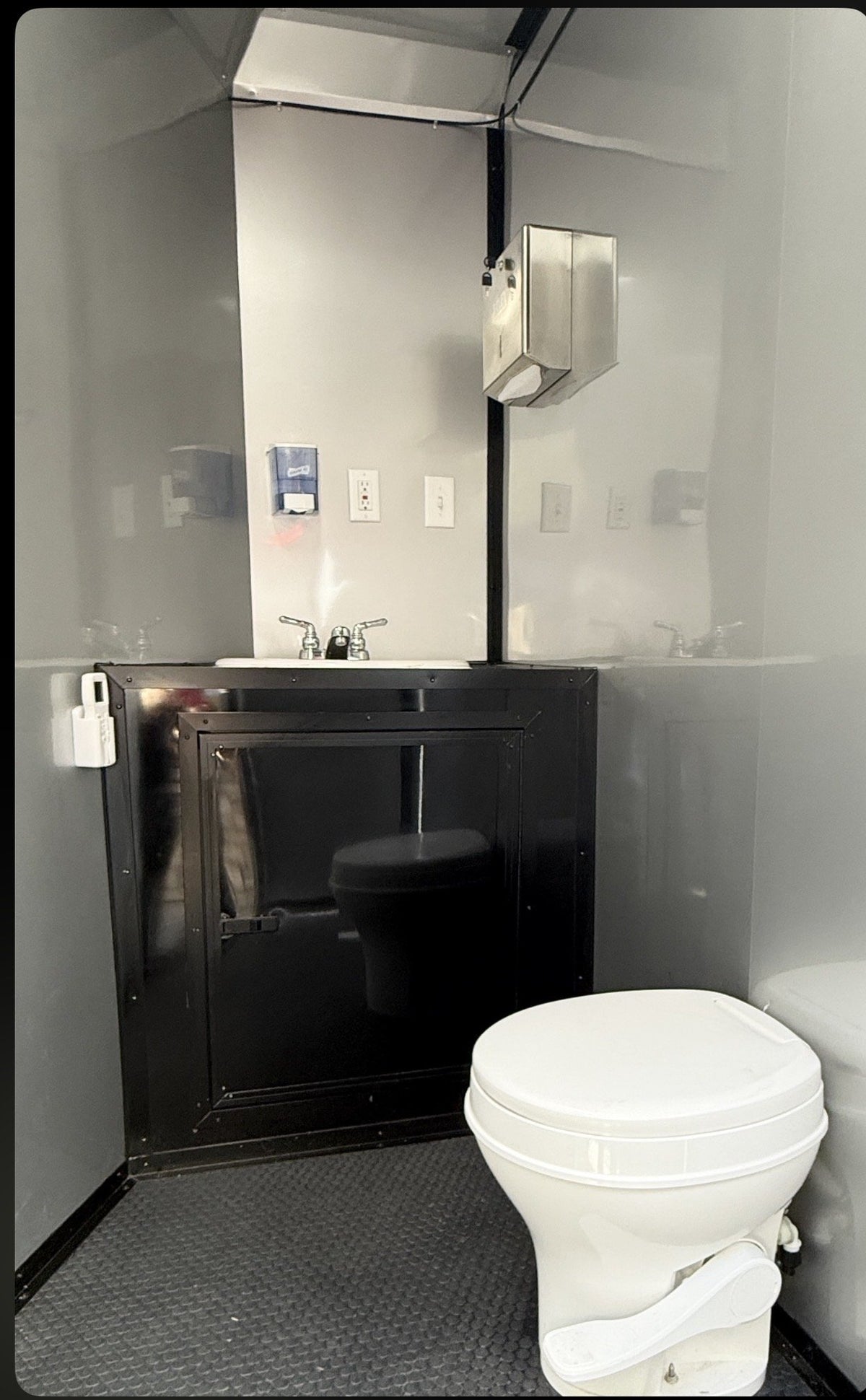 A compact bathroom interior with a white toilet in the foreground, a black vanity cabinet with a sink and faucet behind it, gray walls, and a dark textured floor.