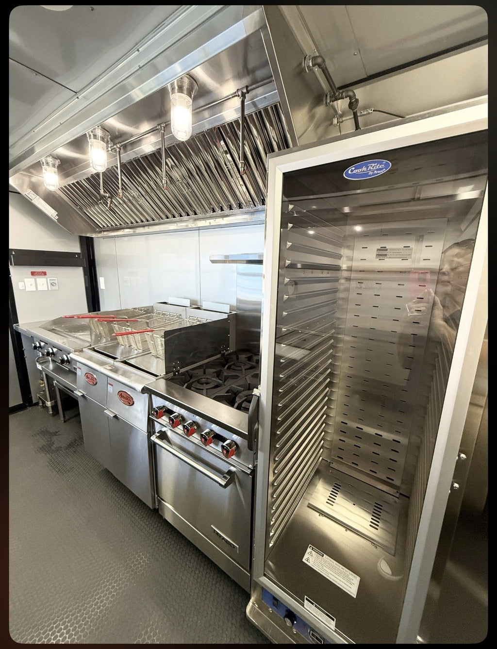 A large stainless steel commercial oven or refrigerator with a glass door stands next to a commercial gas range and fryer in a kitchen setting.