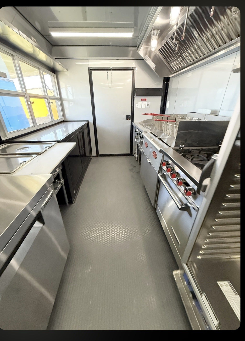 A commercial kitchen interior with stainless steel counters, a large range, a ventilation hood, and a gray textured floor. No people or animals are present.