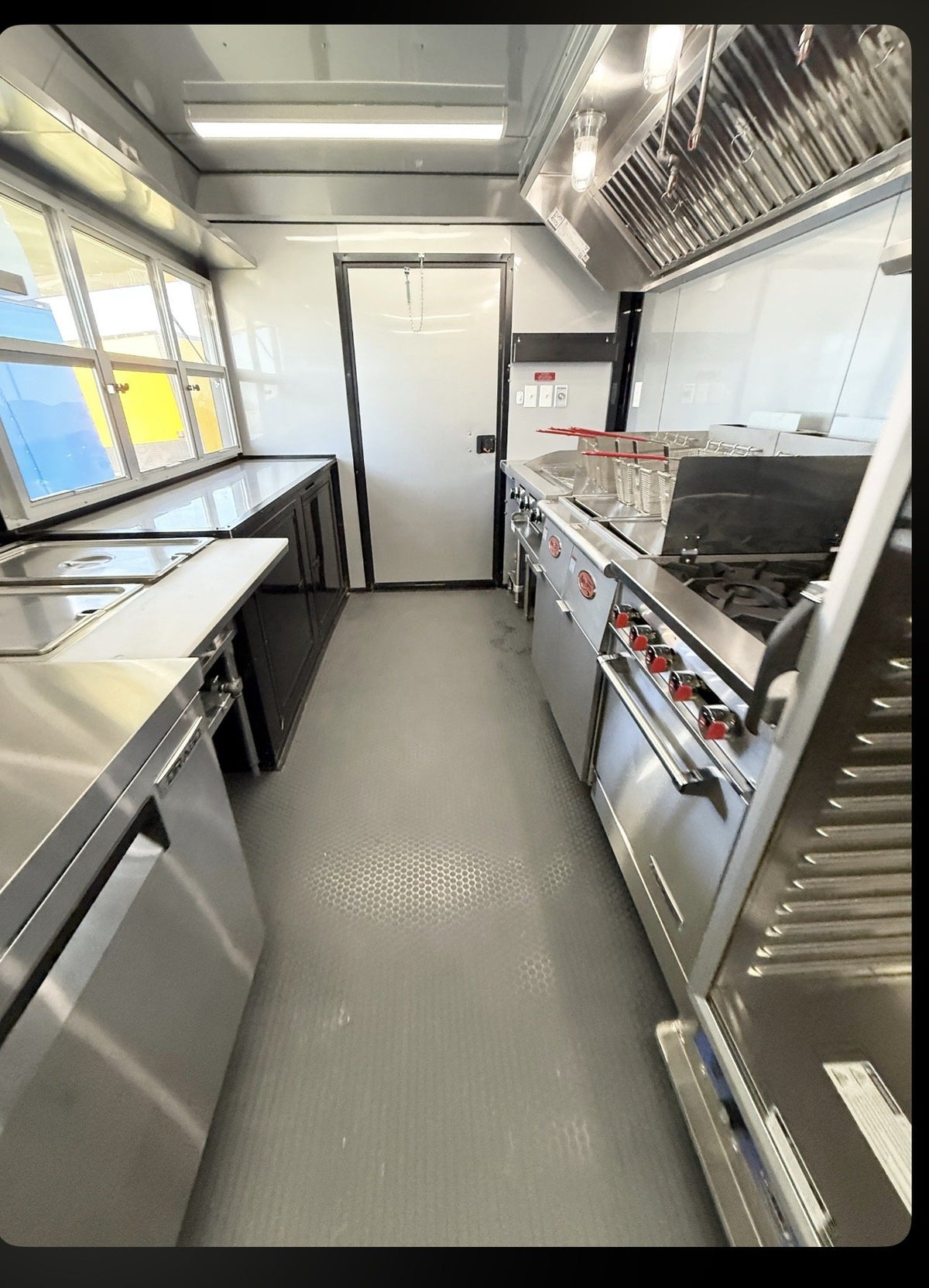A commercial kitchen interior with stainless steel counters, a large range, a ventilation hood, and a gray textured floor. No people or animals are present.