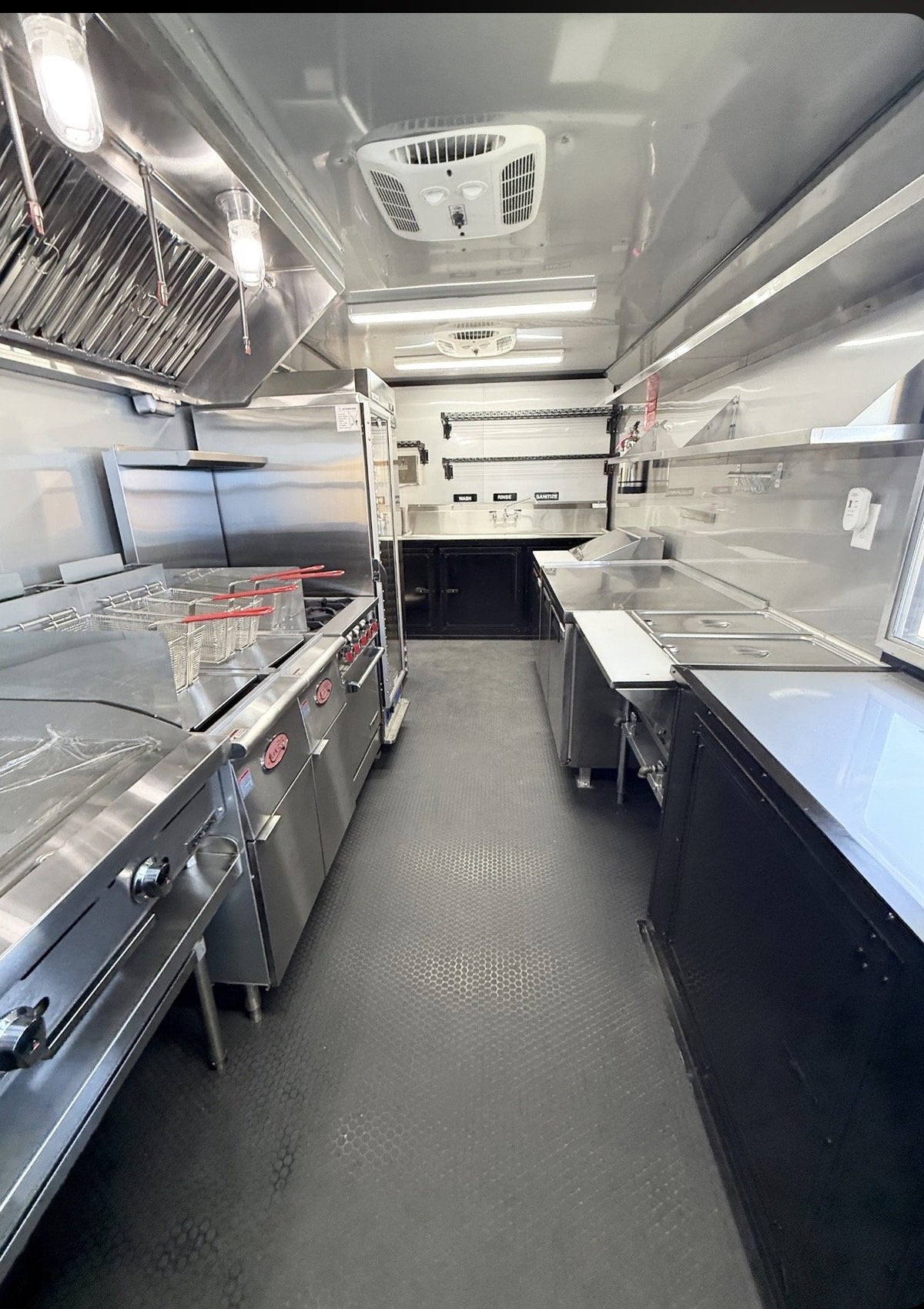 A commercial kitchen interior with stainless steel counters, cooking equipment, shelving, and a gray textured floor, shown from a long aisle perspective.