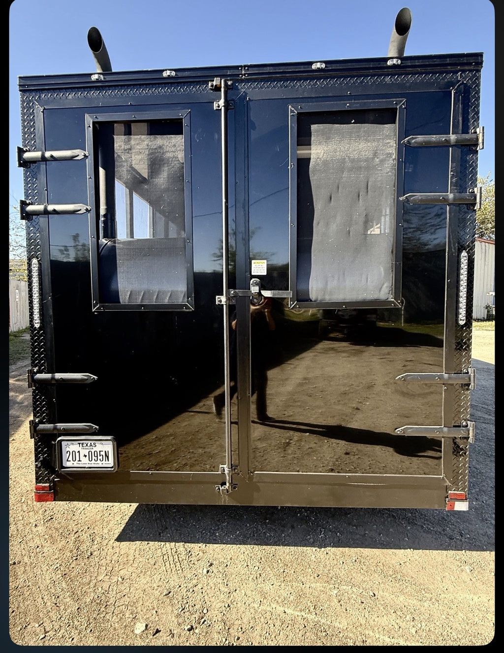 A black metal trailer or cargo container shown from the rear, with two large doors, mesh window panels, side latches, and a Texas license plate, photographed outdoors on a dirt lot under a blue sky.