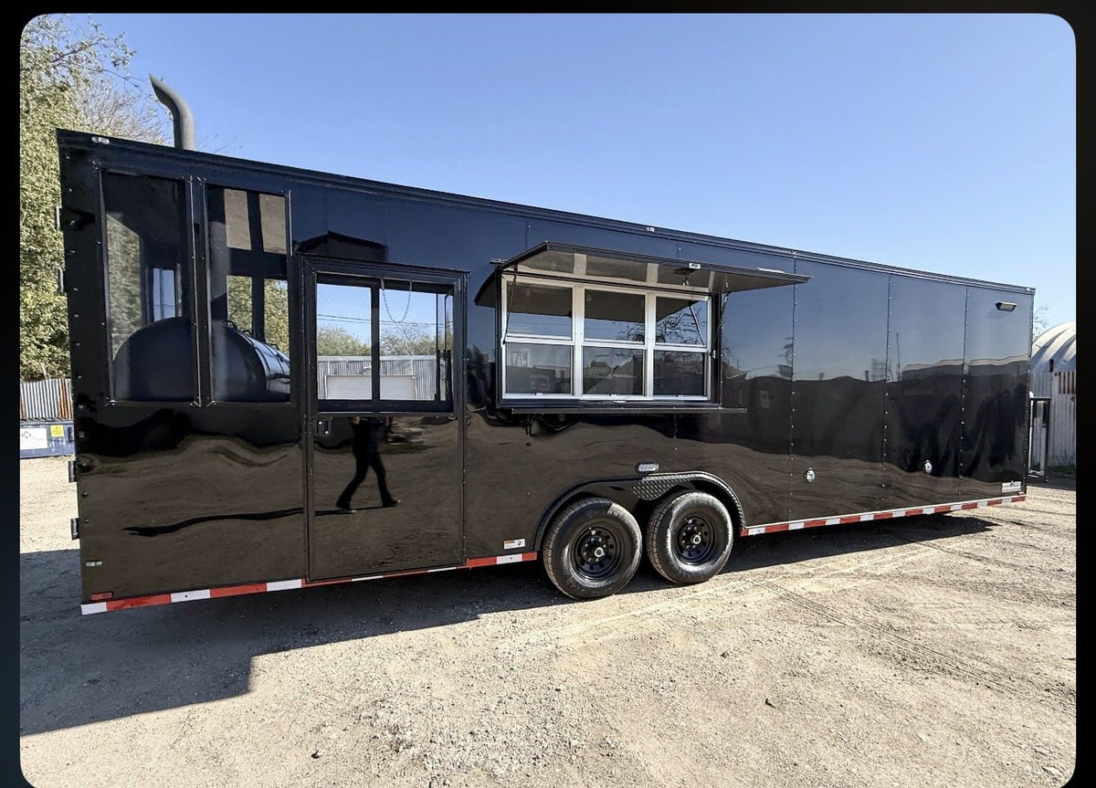 A glossy black food truck trailer parked outdoors on a gravel lot under a clear blue sky. The trailer has two rear wheels, side windows, and an open serving window with a grill visible above it.
