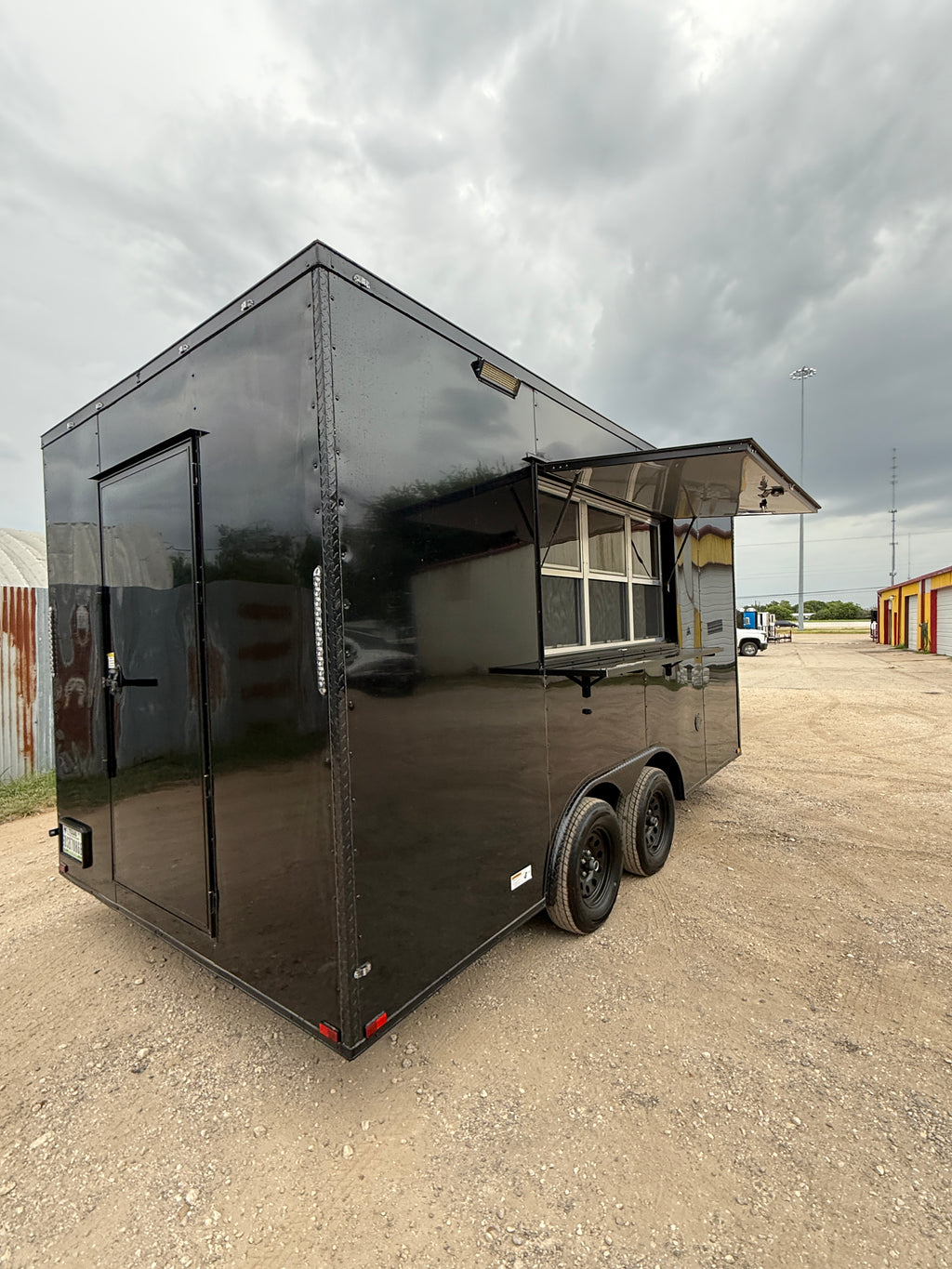 16 FT black blackout cooking trailer, front three-quarter view under a cloudy sky with the serving window awning raised