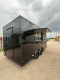 16 FT black blackout cooking trailer, front three-quarter view under a cloudy sky with the serving window awning raised